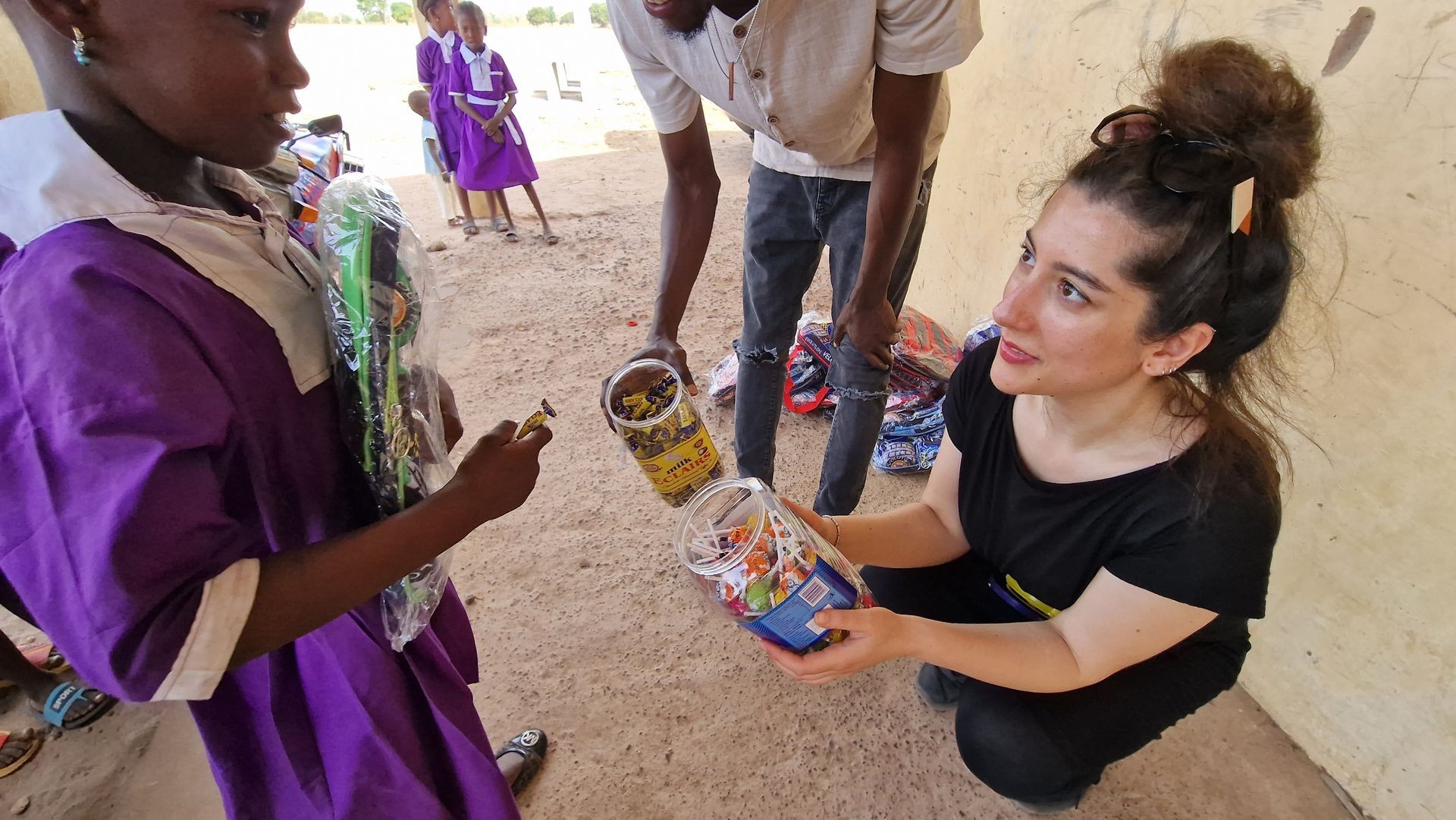 Woman handing out candies to a young girl in a school uniform outdoors.